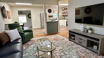 Una sala de estar con un sofá, una mesa de centro y un televisor. Se observa una cocina al fondo.