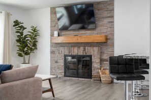 Ein modernes Wohnzimmer mit Kamin aus Stein, einem flachbildfernseher an der Wand, einer Sitzecke mit Sofa und Sesseln sowie einem Couchtisch.