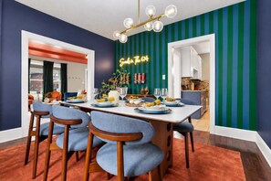 A dining area with a long table set for four, chairs with blue upholstery, a chandelier with multiple globes, and a neon sign on the wall. - The 614 Club Pool Hot Tub Fire Pit Games (Columbus)