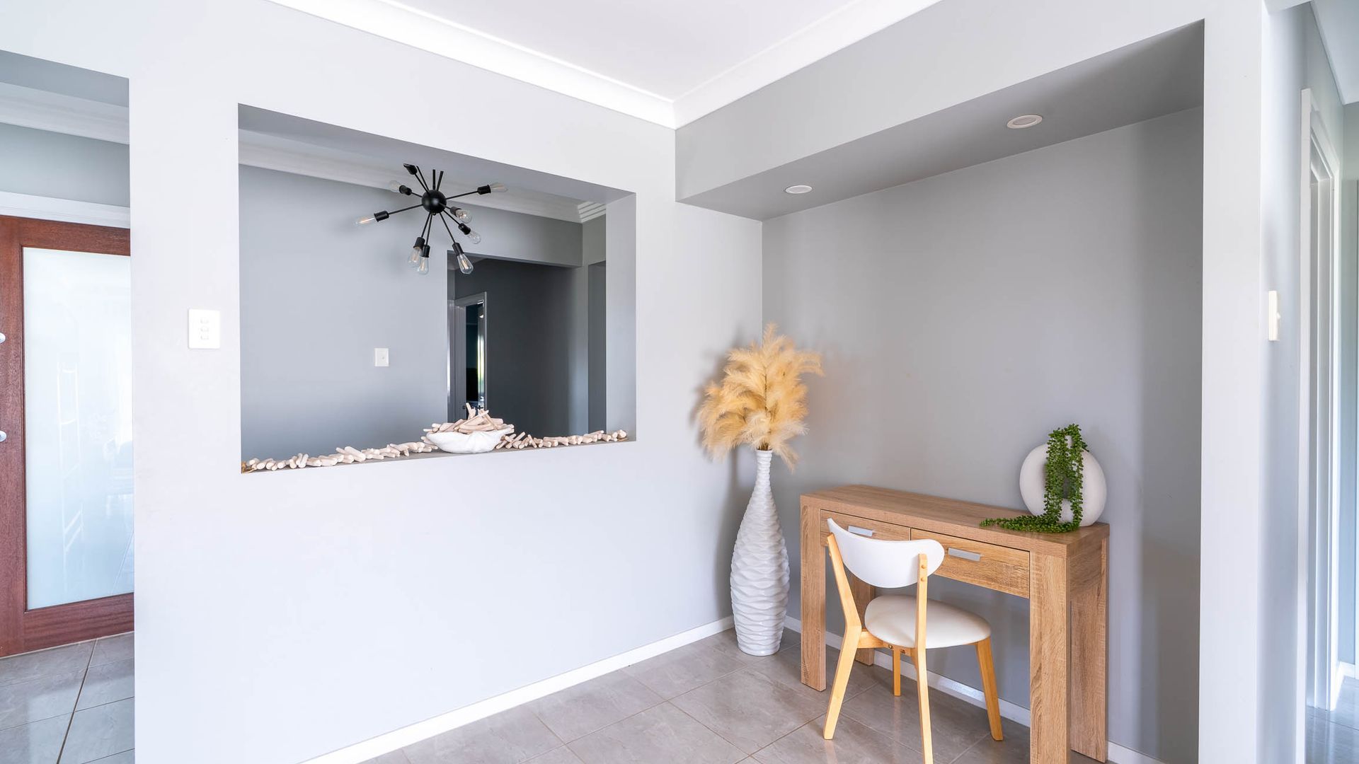 Un interior moderno con una consola de madera, una silla blanca, un espejo decorativo y un jarrón con plumas de pájaro.
