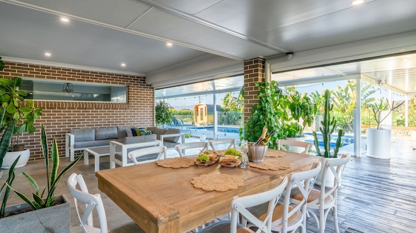 An outdoor dining area with a brick wall, white furniture, and a pool in the background. - Hillside Residence ~ Picturesque mountain views with four spacious bedrooms (Nowra Hill)