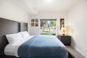 Ein Schlafzimmer mit einem Bett, einem Nachttisch, einer Lampe und einem Fenster mit Blick auf einen Garten.