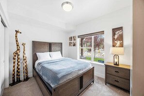 Quarto com cama, mesa de cabeceira, abajur, janela e decoração com girafas.