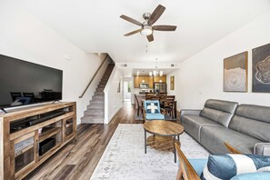 Ein modernes Wohnzimmer mit Flachbildfernseher, grauem Sofa, einer Holz-Unterhaltungsecke und einem Essbereich mit Holzsesseln und einer Pendelleuchte.