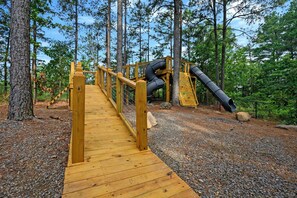 Eine Holzbrücke führt zu einem Spielplatz mit Kletterwänden und einer Rutsche.