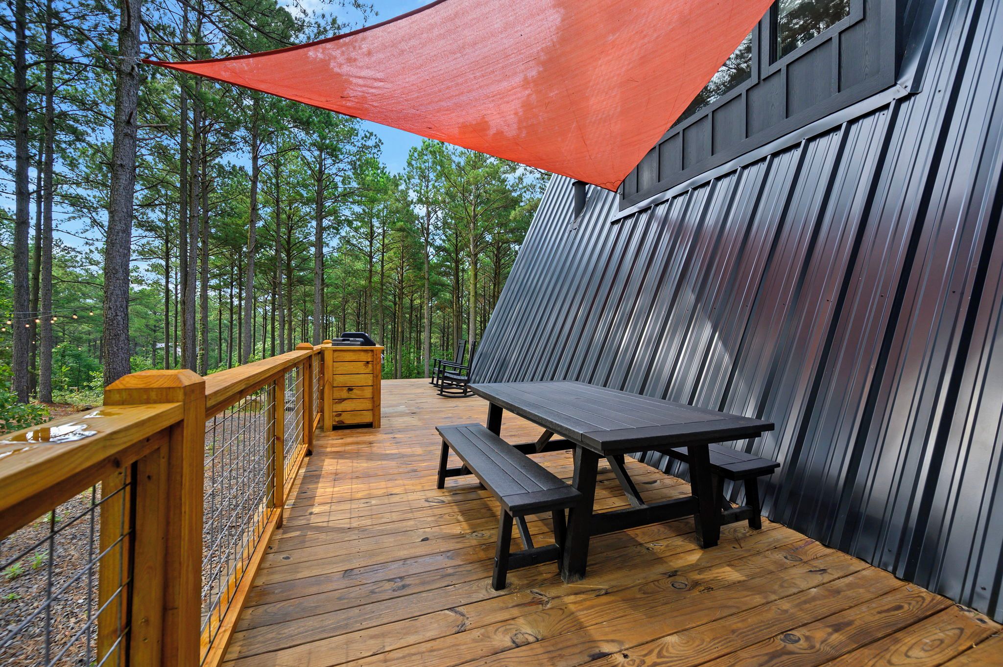 A wooden deck with a picnic table and benches, surrounded by a metal fence and a red shade sail.
