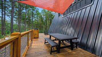 A wooden deck with a picnic table and benches, surrounded by a metal fence and a red shade sail.