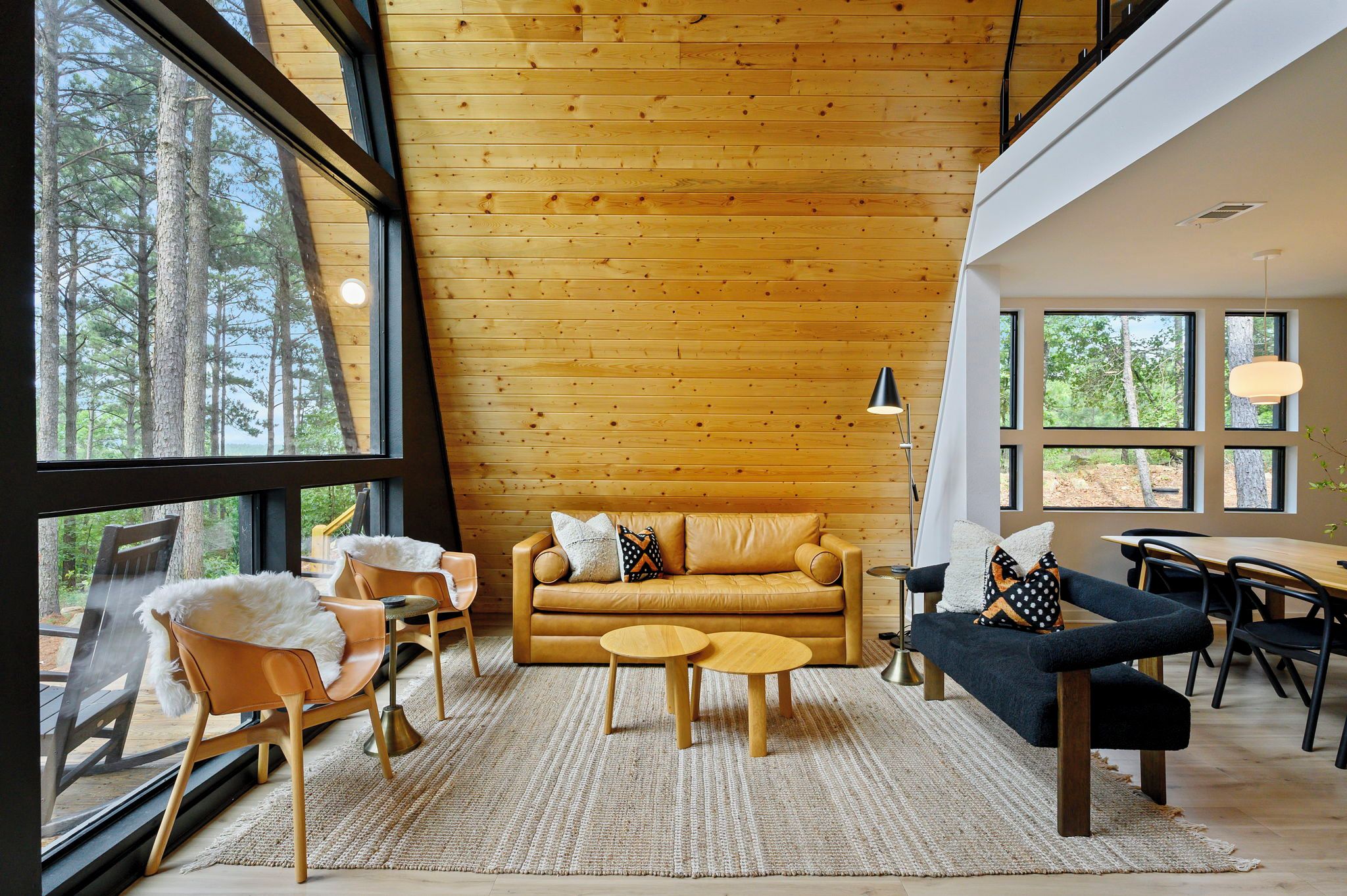 A cozy cabin interior with a wooden ceiling, a leather sofa, two armchairs, a coffee table, and a dining table with chairs.