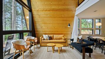 A cozy cabin interior with a wooden ceiling, a leather sofa, two armchairs, a coffee table, and a dining table with chairs.