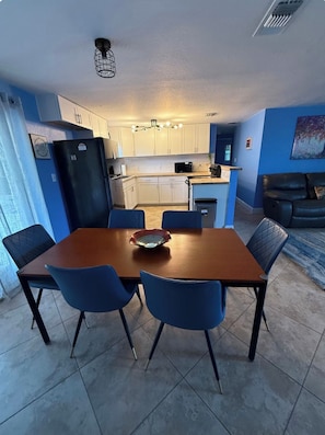 Ein Essbereich mit einem Holztisch und blauen Stühlen, einem schwarzen Kühlschrank sowie einer Küche im Hintergrund.