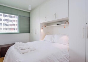 Ein Hotelzimmer mit einem Bett, weißer Bettwäsche, einem Nachttisch aus Holz, einem Koffer und einem Fenster mit Jalousien.