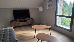 Een moderne woonkamer met een flatscreen televisie, houten meubels, een groot raam en een lamp.