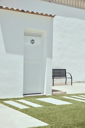 A white building with a door featuring a male and female restroom symbol, a bench, and a grassy area with a concrete path.
