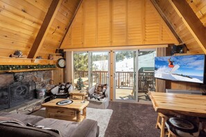 Ein gemütliches Blockhaus-Interieur mit Kamin, Fernseher und einer Terrasse mit Blick auf eine verschneite Landschaft.
