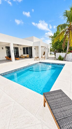 A rectangular swimming pool with clear blue water, surrounded by a white tiled deck. There are lounge chairs and a covered seating area with a glass door. - Newly Listed -Canal front 2-bedroom Villa (Providenciales)