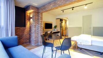 Un salon moderne avec un mur d’accent en briques, un téléviseur à écran plat, un coin repas avec une table et des chaises, et un lit recouvert d’une couette blanche.