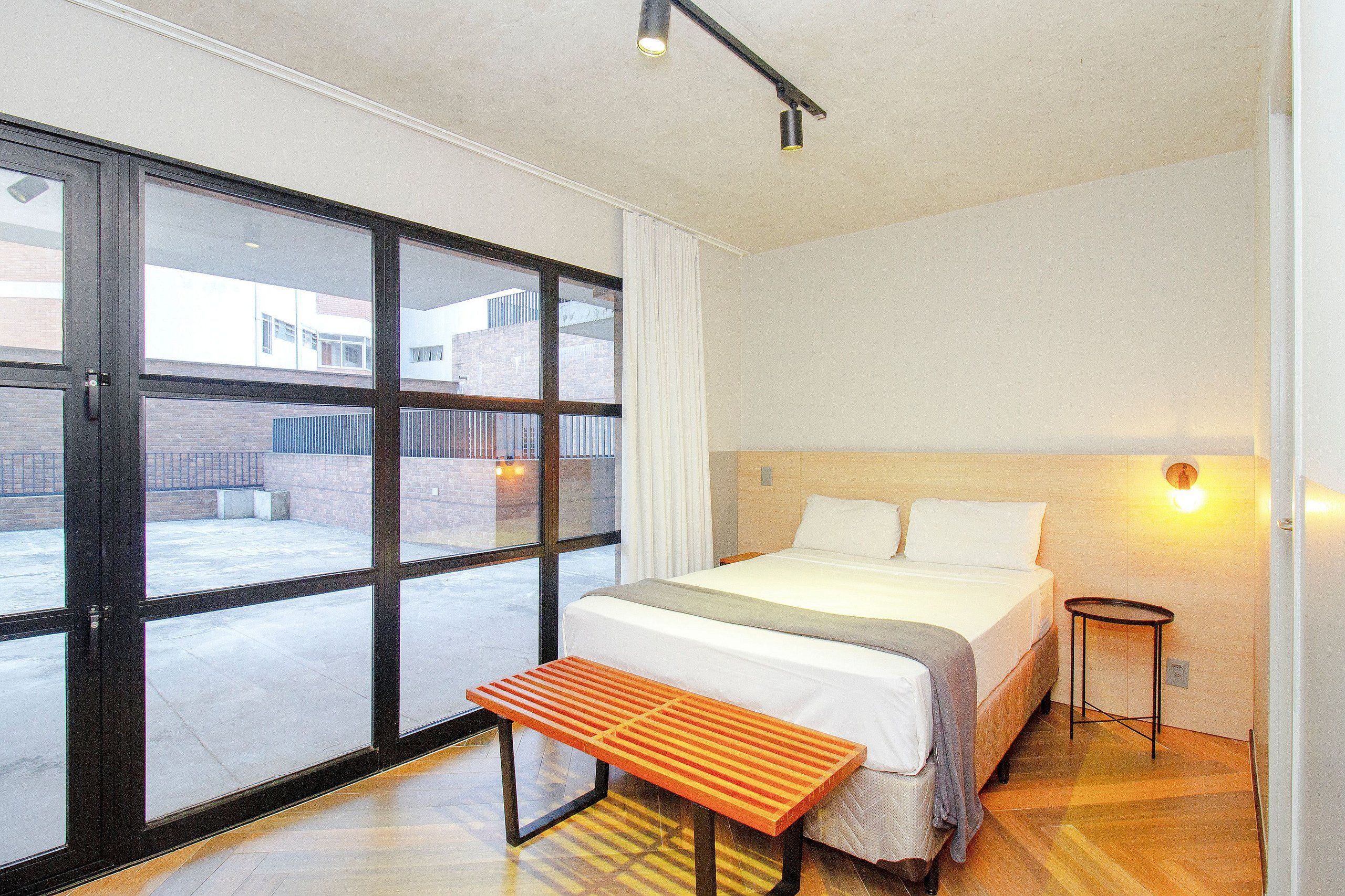 Une chambre d’hôtel avec un lit, un banc, une petite table, une table de chevet et une fenêtre donnant sur l’extérieur.