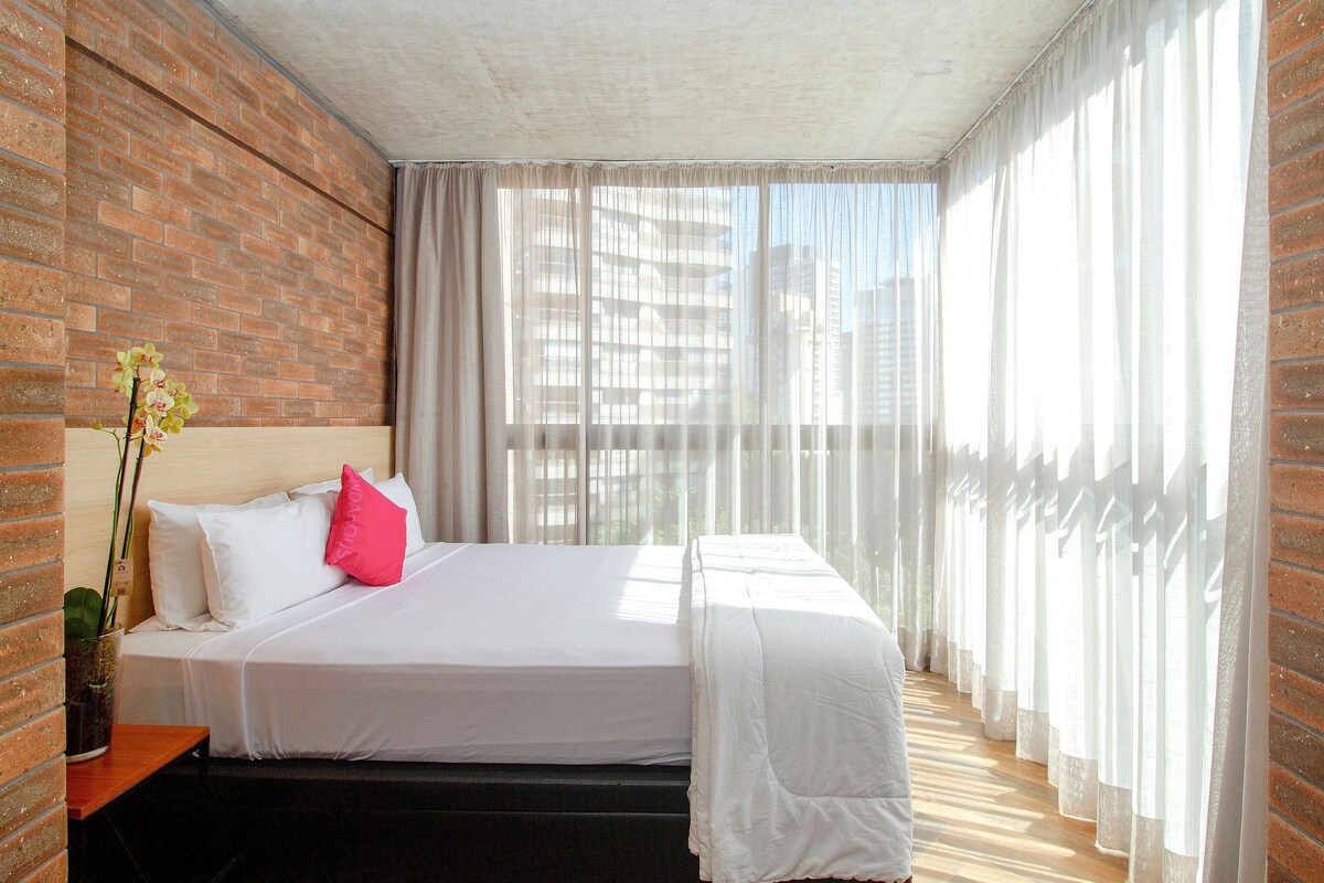 A hotel room with a brick wall, a bed with white bedding, a pink pillow, a bedside table with a vase of flowers, and sheer curtains.