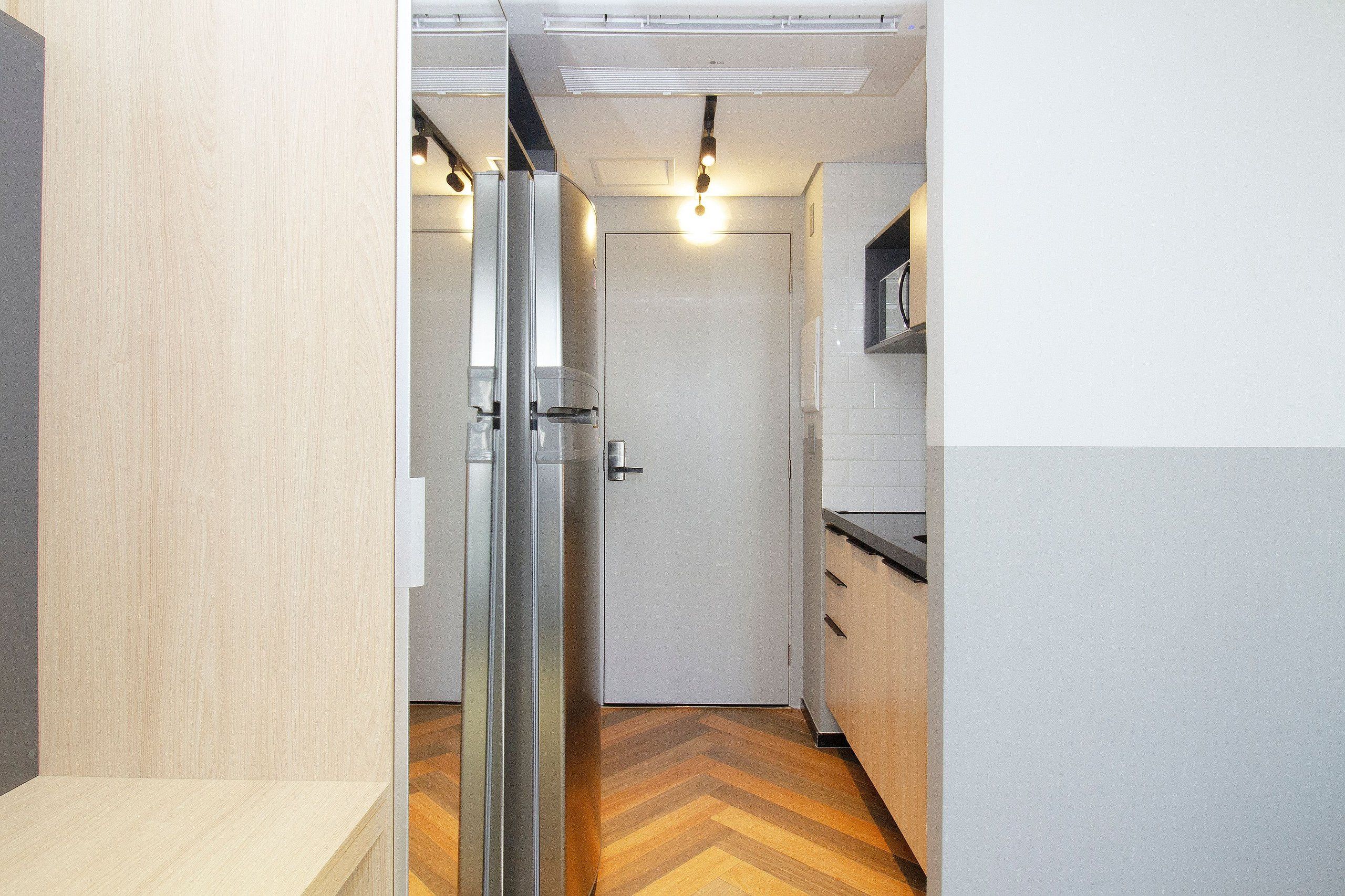 Un couloir étroit avec une porte en verre donnant sur une pièce, une porte blanche au fond, et un coin cuisine avec un plan de travail et des placards.
