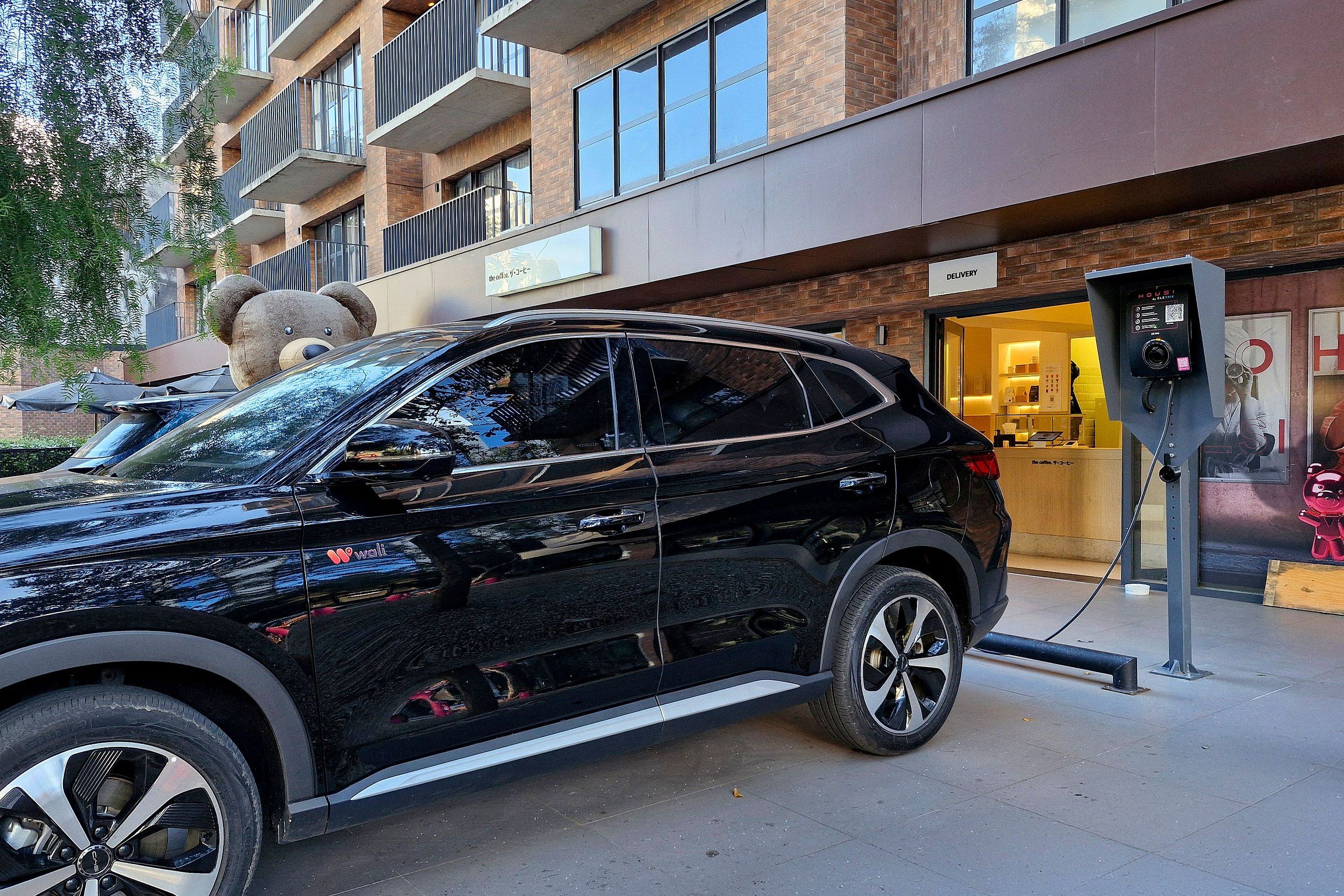 A black SUV parked at a charging station in front of a building with a sign that reads 'DELIVERY'.