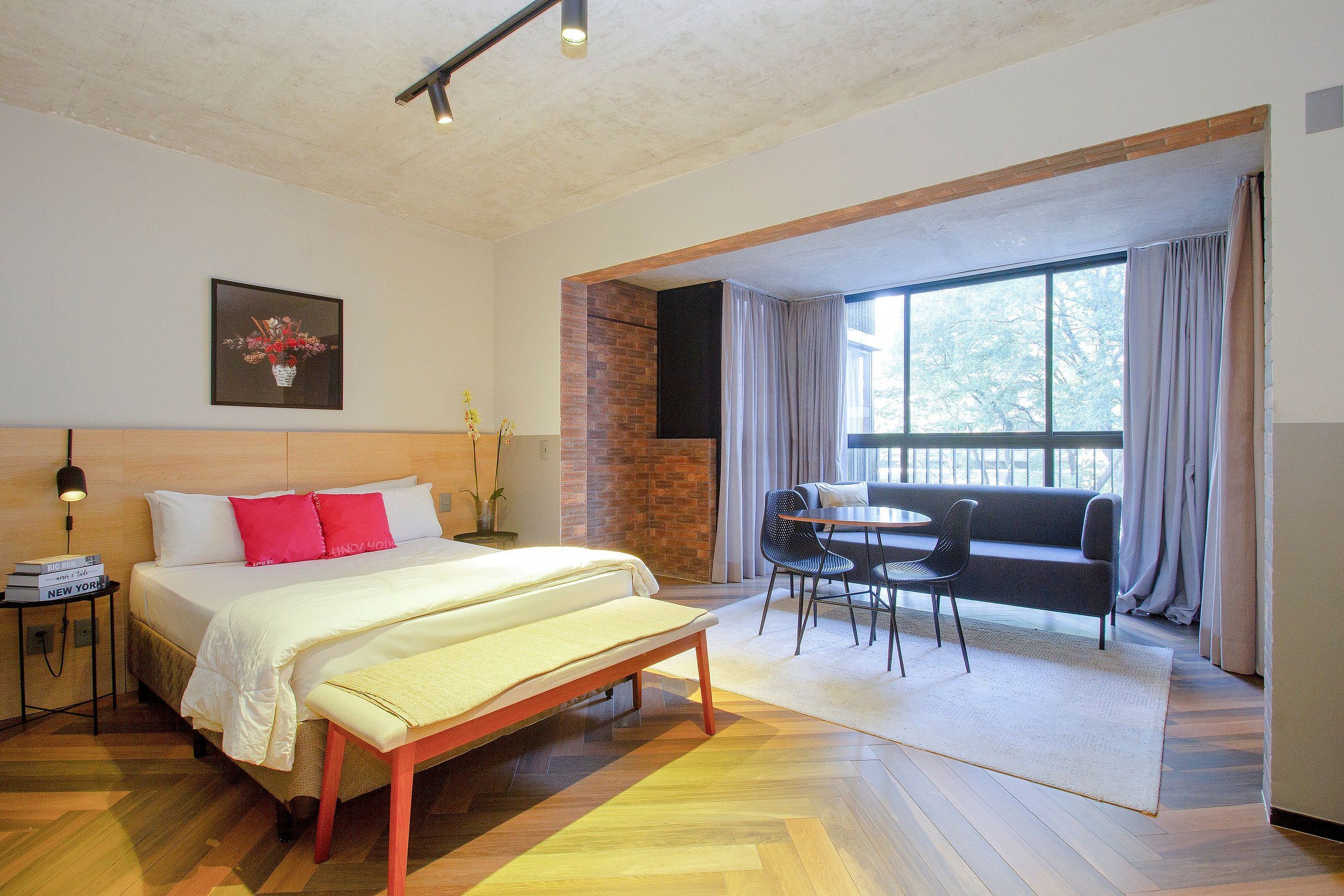 Una habitación de hotel con cama, mesita de noche, banco y un área de descanso con mesa y sillas.