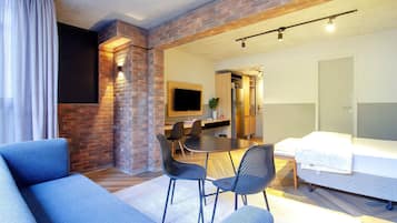 Un salon moderne avec un mur d’accent en briques, un téléviseur à écran plat, un coin repas avec une table et des chaises, et un lit recouvert d’une couette blanche.