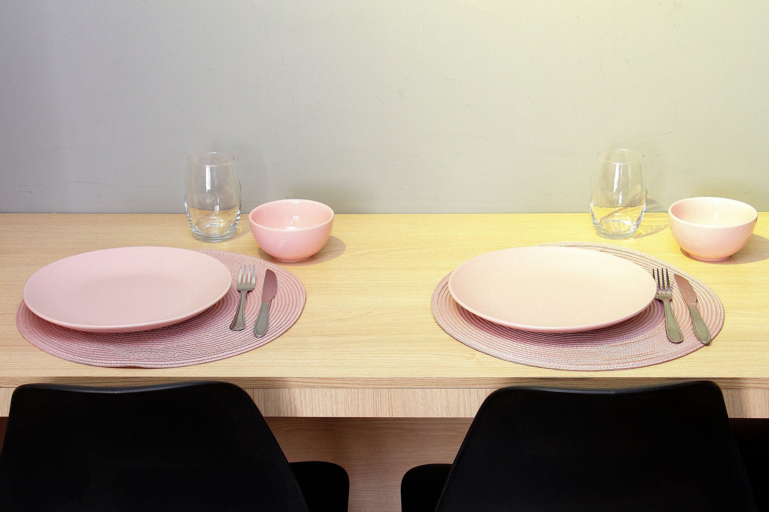 Mesa de jantar posta para dois, com pratos cor-de-rosa, talheres e copos.