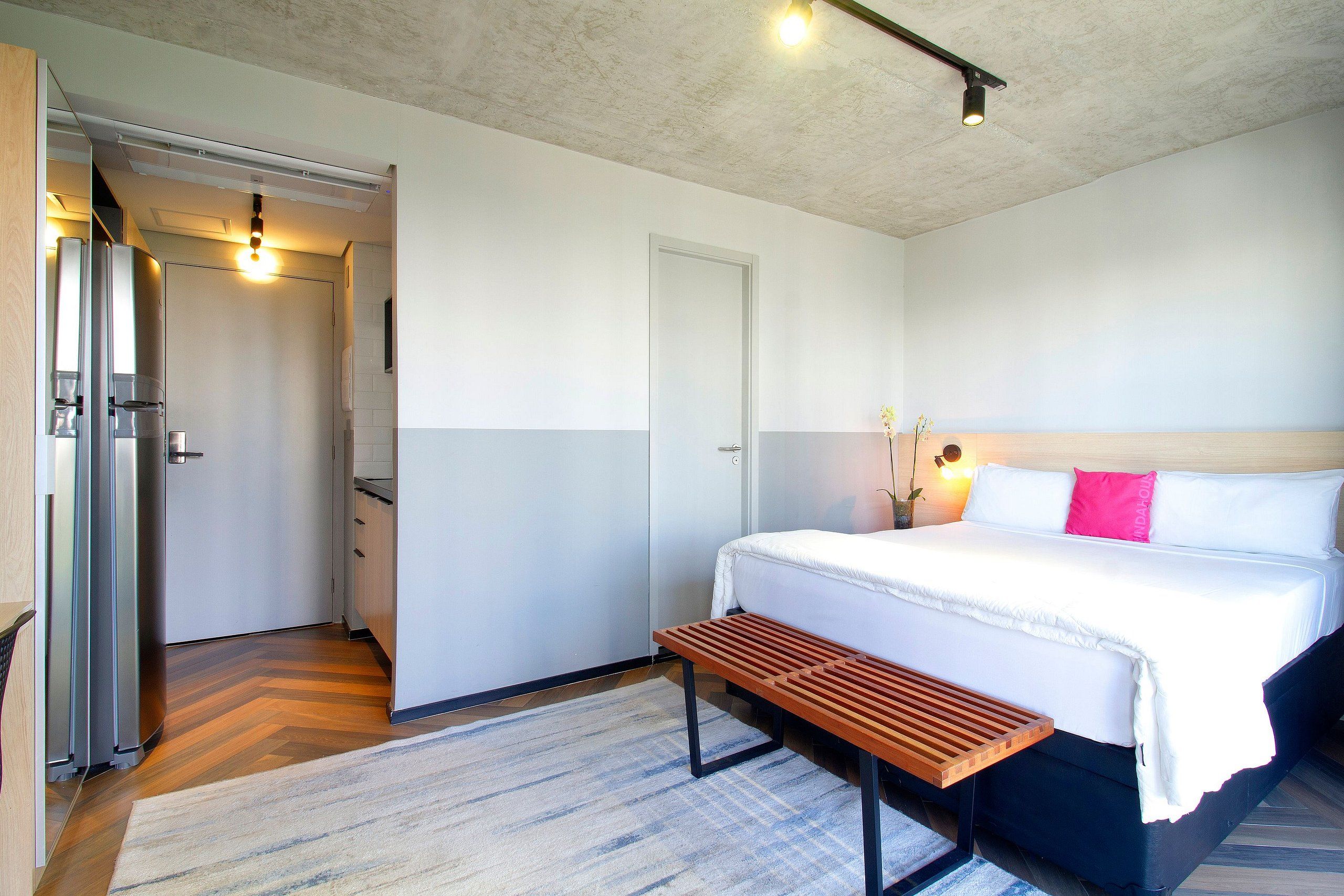 A hotel room with a bed, bedside table, bench, and a small bedside plant.