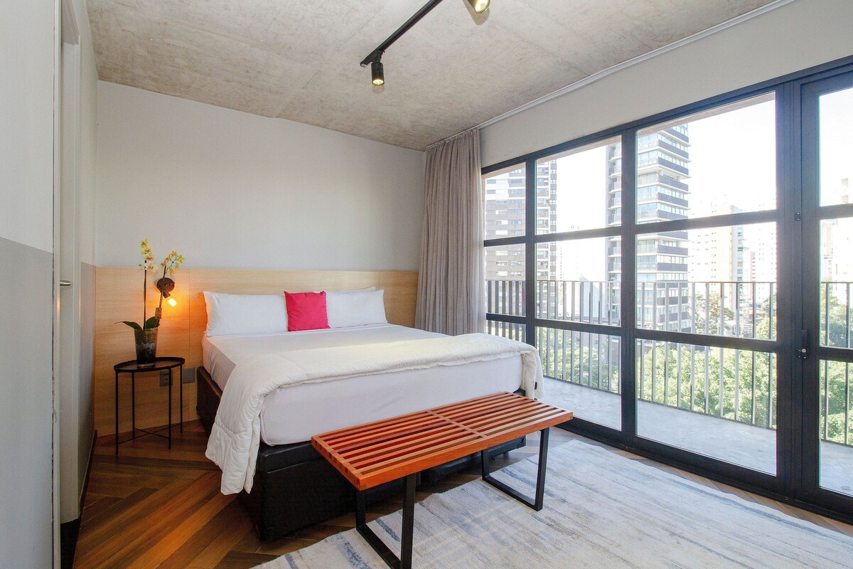 A hotel room with a large bed, a bedside table with a lamp, a bench, and a view of buildings through large windows.