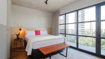 Habitación de hotel con una cama grande, una mesita de noche con lámpara, un banco y vistas a edificios a través de amplias ventanas.