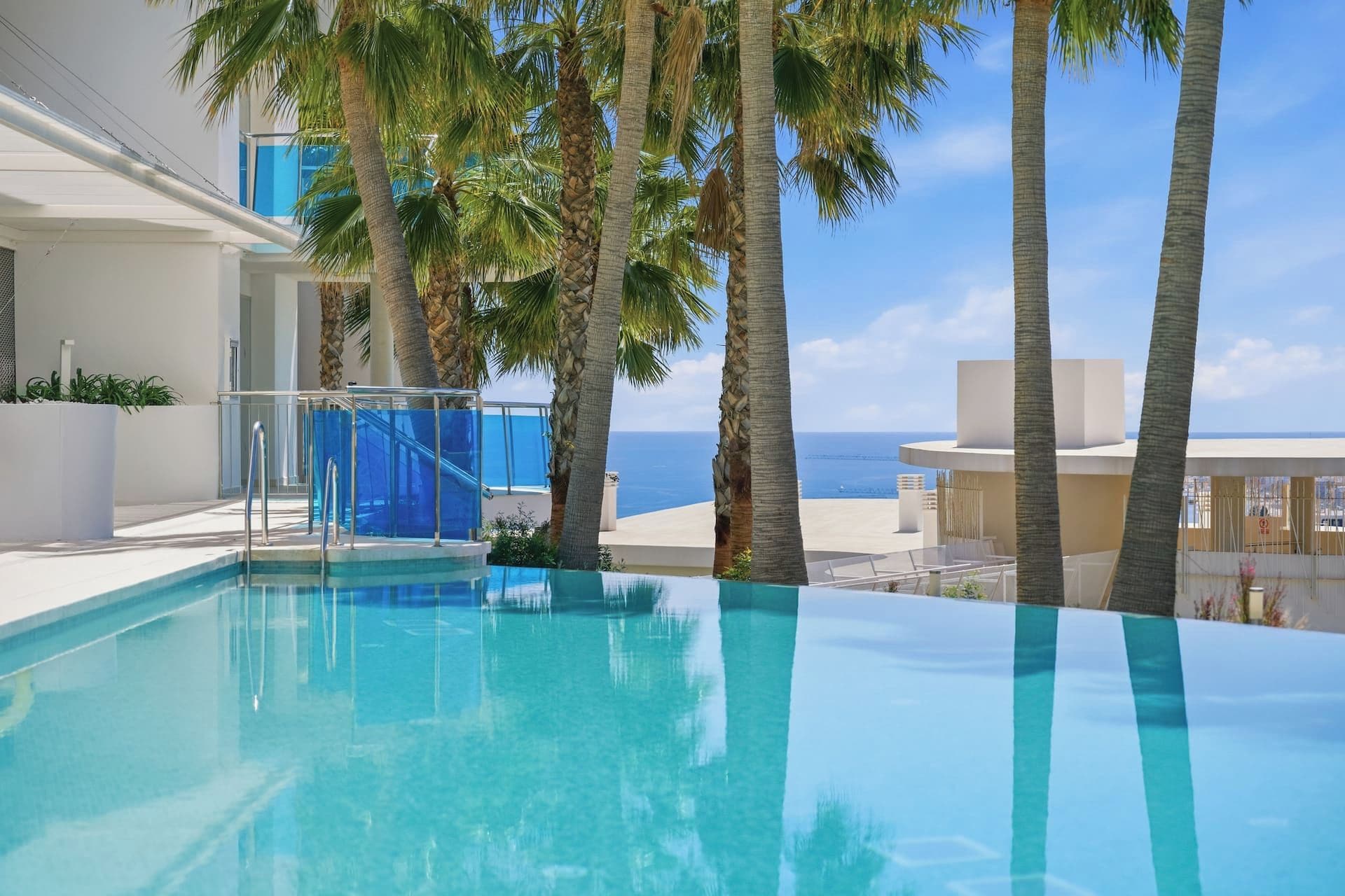 A swimming pool with clear blue water, surrounded by palm trees and a view of the sea.