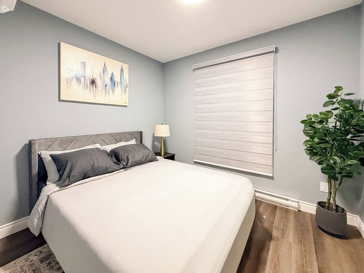 A bedroom with a bed, a nightstand, a lamp, a window with blinds, and a potted plant.