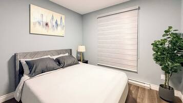 A bedroom with a bed, a nightstand, a lamp, a window with blinds, and a potted plant.