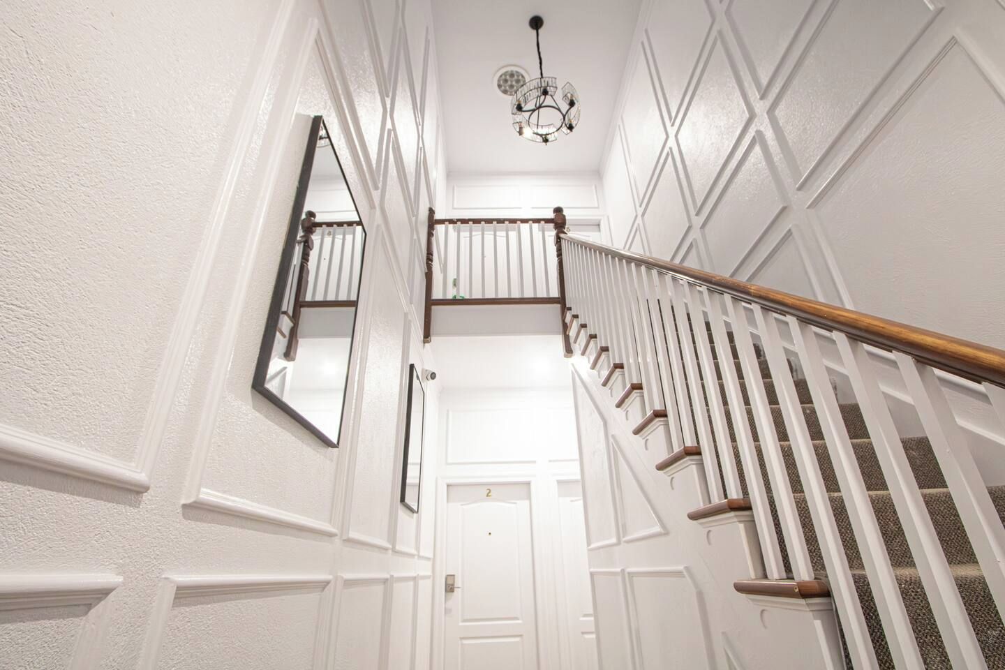 A white interior hallway with a wooden staircase, a chandelier, and a door with the number '2' on it.