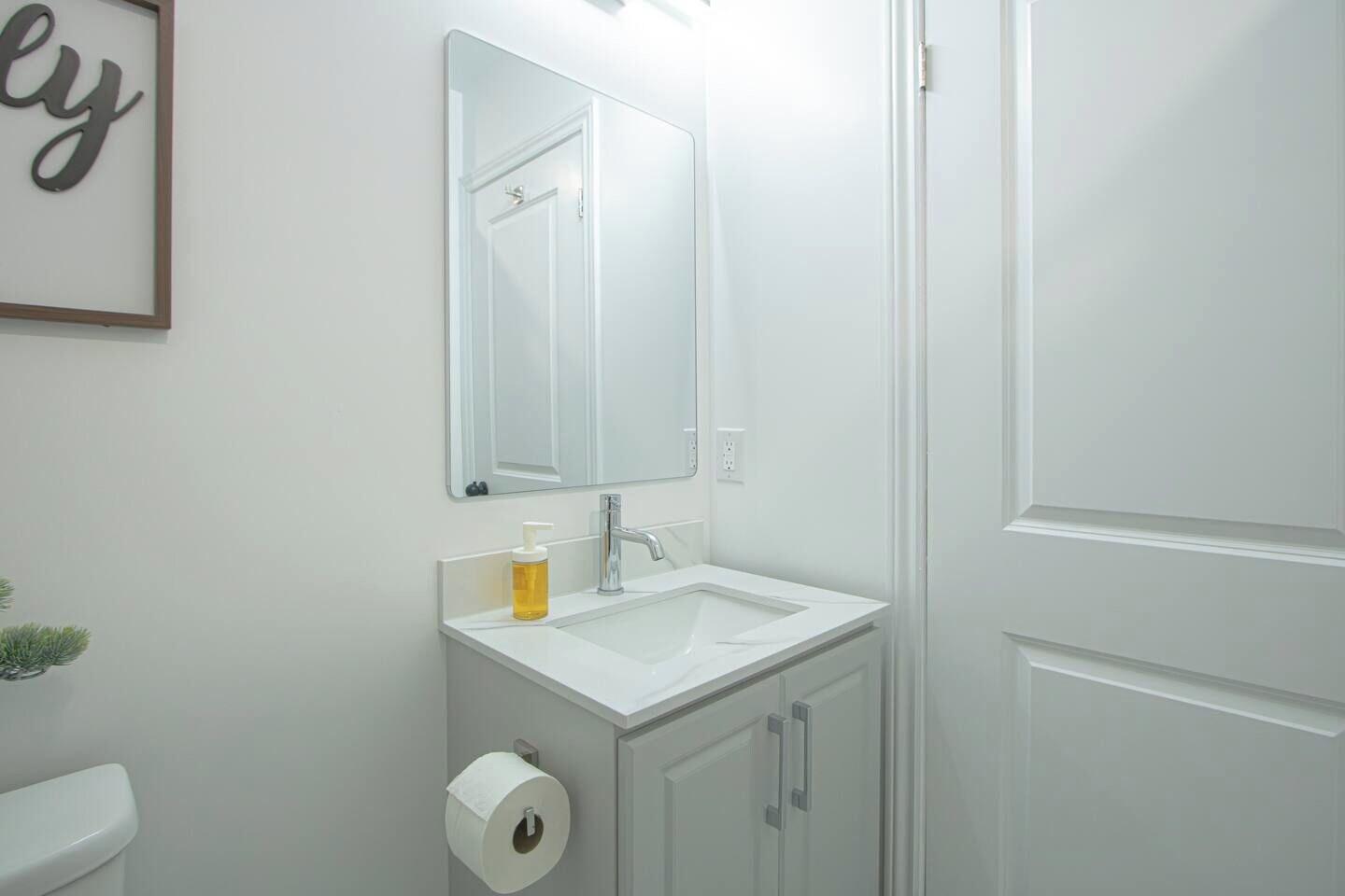 Un baño con lavamanos blanco, un espejo, un inodoro y un cartel enmarcado en la pared.