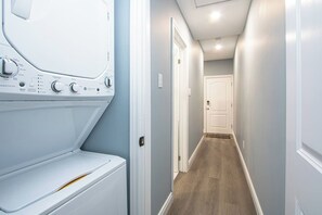 Une buanderie équipée d’un lave-linge et d’un sèche-linge à chargement frontal, un couloir avec un plancher en bois et une porte au bout.