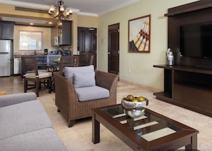A living room with a sofa, a coffee table with a bowl of fruit, a television, and a kitchen area in the background.