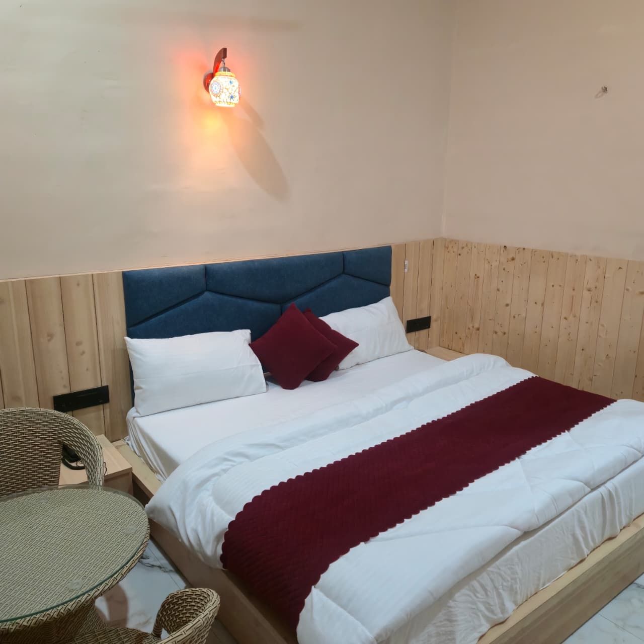 Un letto rifatto con lenzuola bianche e bordeaux, una testiera imbottita blu e un punto luce fissato al muro.