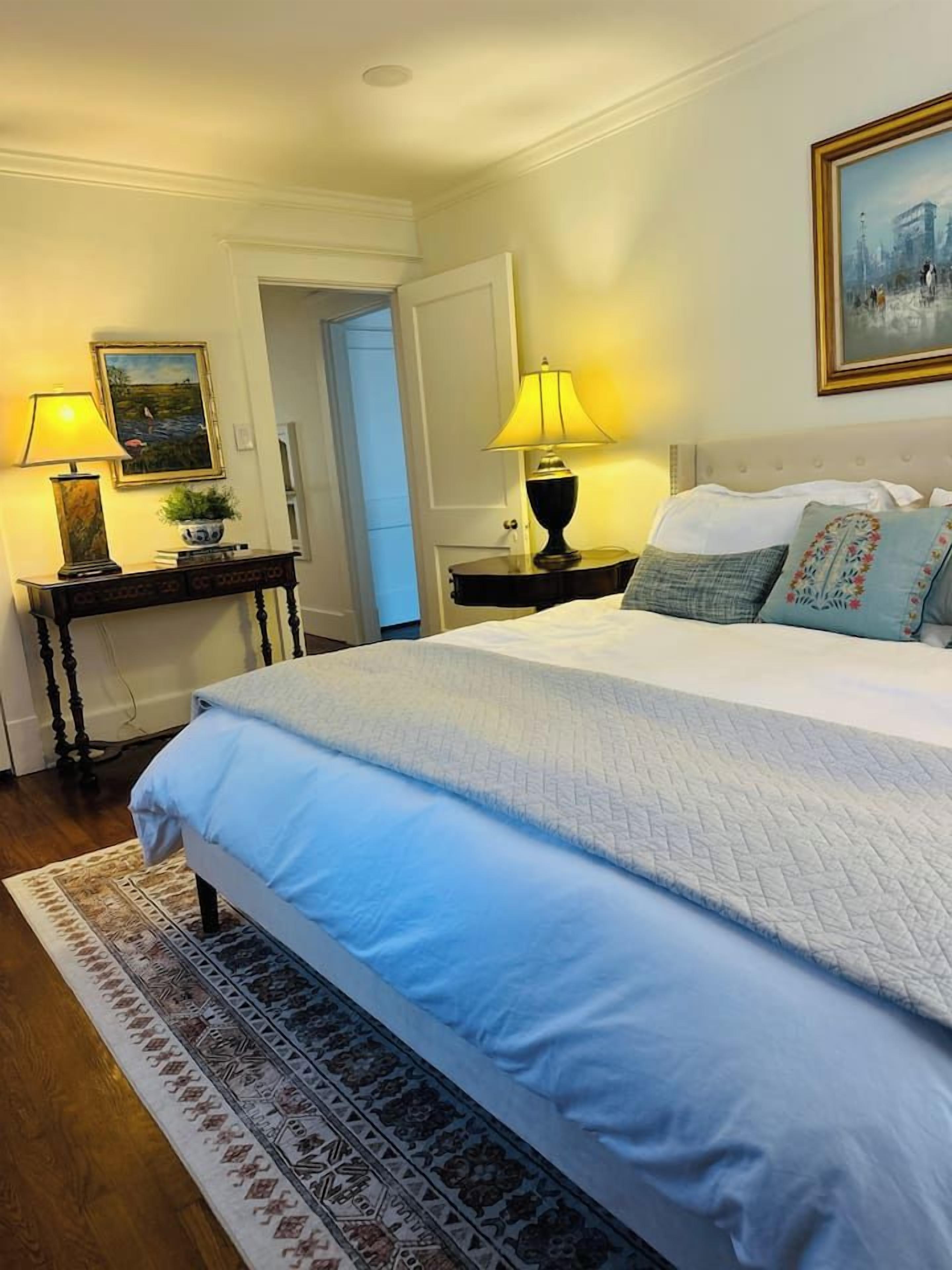 Une chambre à coucher avec un grand lit, deux tables de chevet avec des lampes, un tableau au mur et une porte donnant sur une autre pièce.