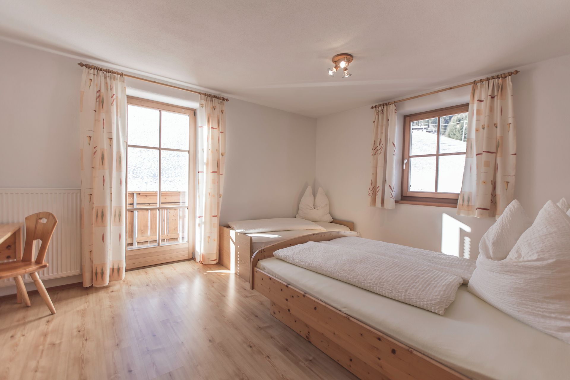 Una habitación con piso de madera, una cama con ropa de cama blanca, una silla y una ventana con cortinas.