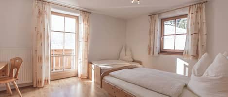 A room with a wooden floor, a bed with white bedding, a chair, and a window with curtains.