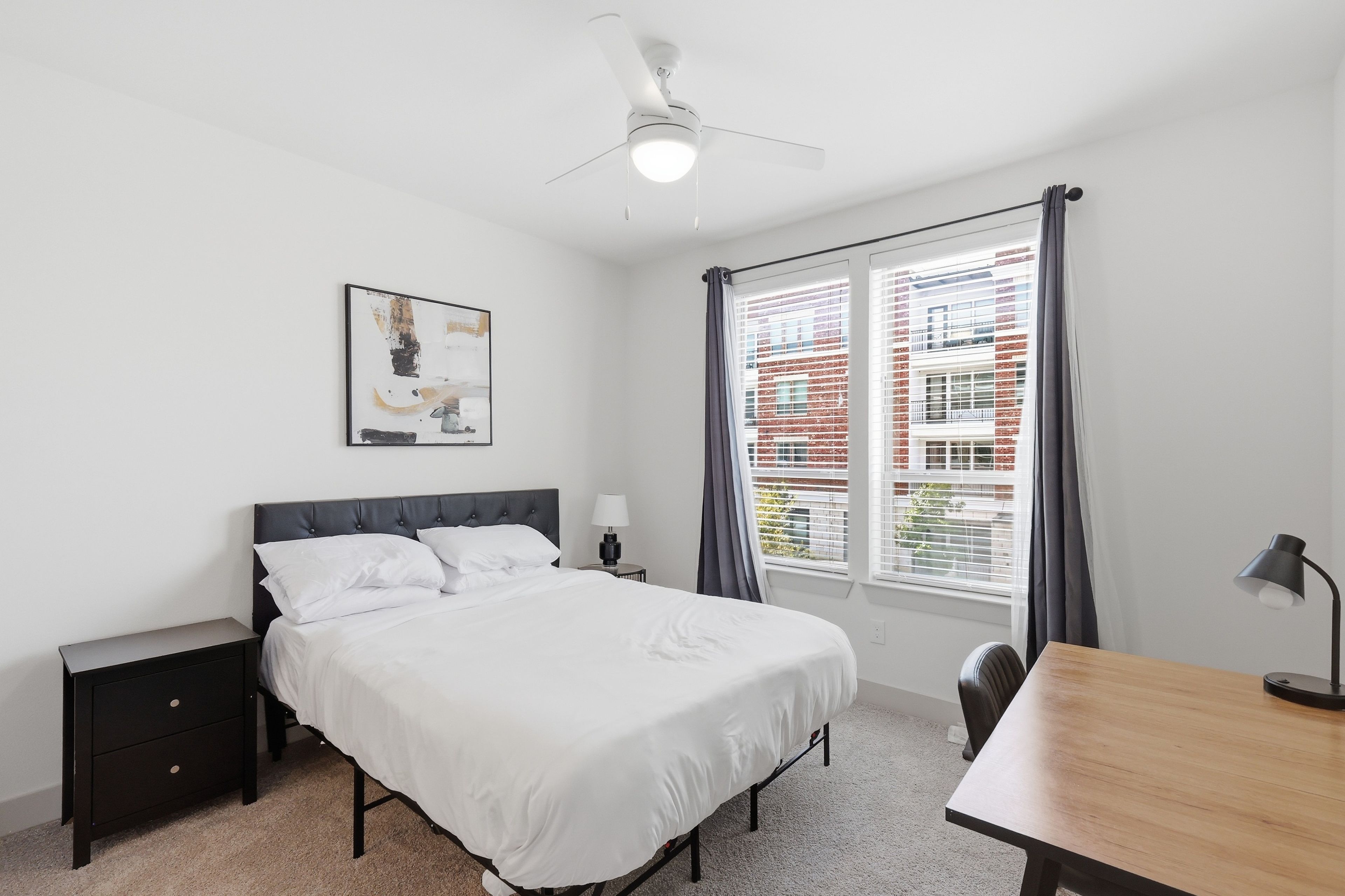 A bedroom with a bed, bedside tables, a desk, a ceiling fan, and a window with curtains.