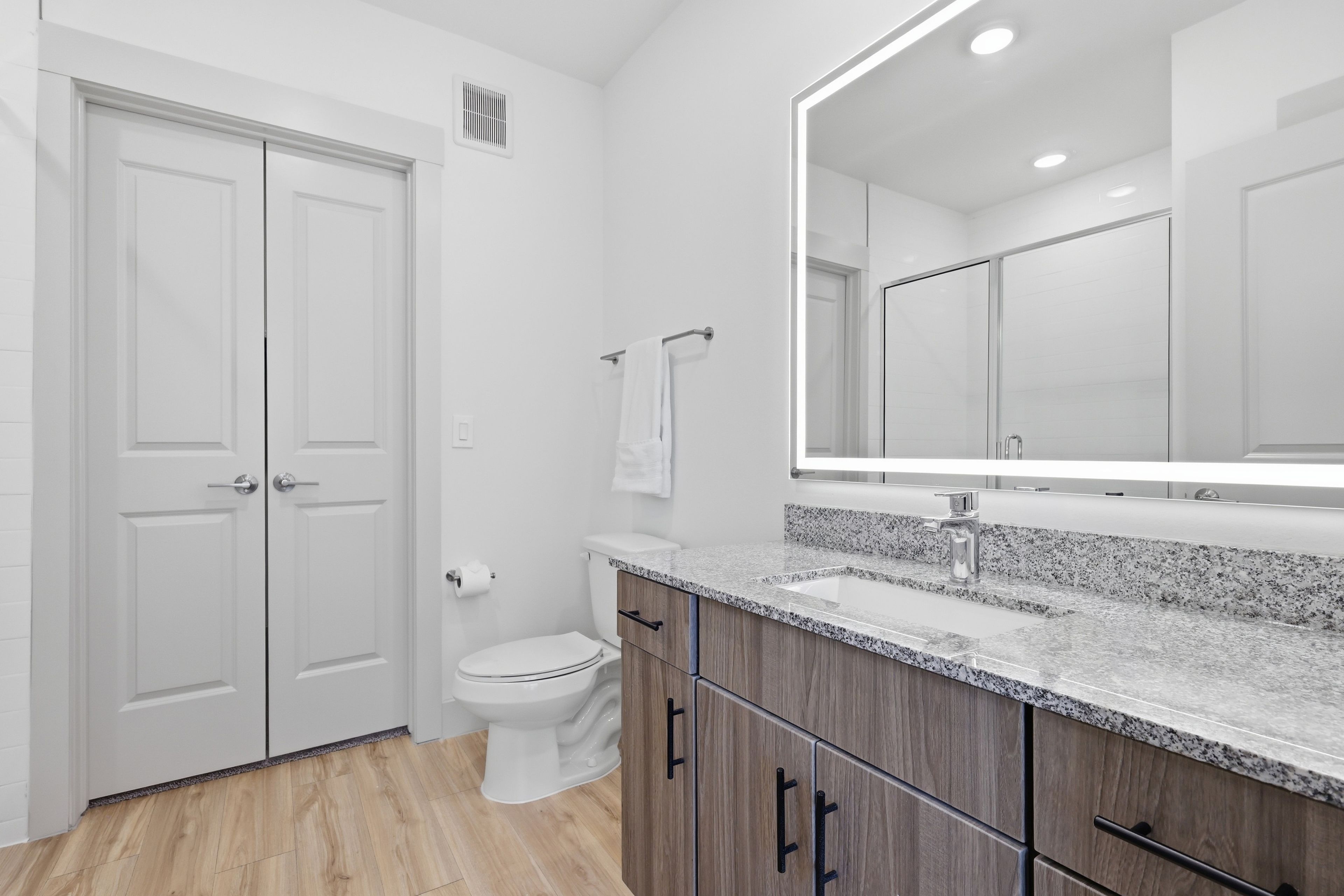 A modern bathroom with a double door, a toilet, a sink with a large mirror, and wooden cabinetry.