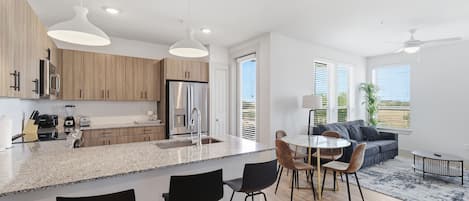 Cozinha moderna com ilha central, banquetas, mesa de jantar e sofá numa sala de estar ampla e iluminada com grandes janelas.