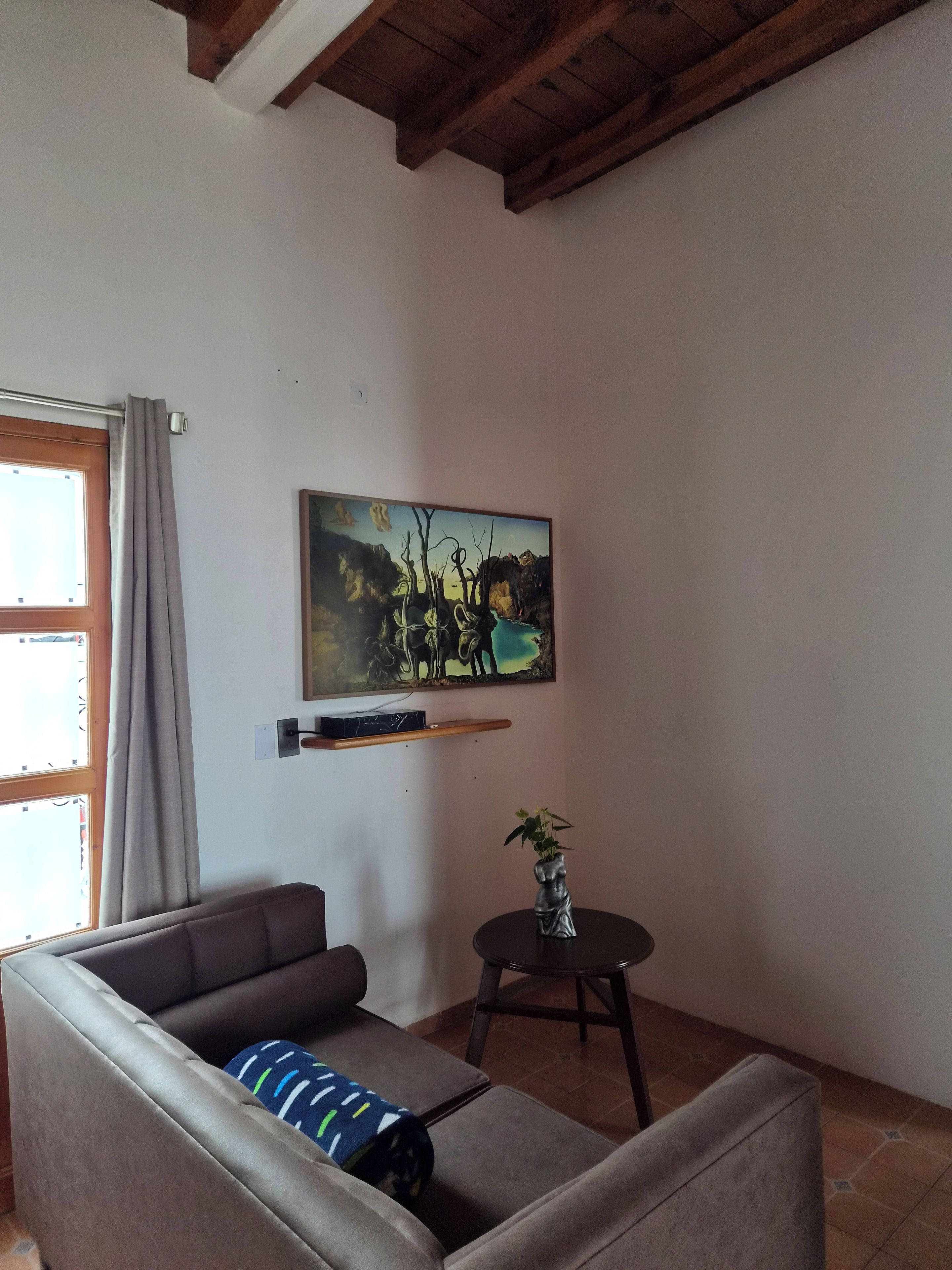 A living room with a sofa, a small table with a plant, a painting on the wall, and a window with curtains.