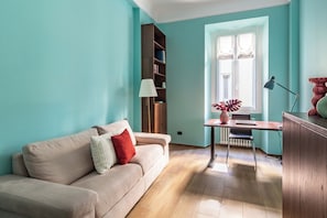 A living room with a beige sofa, a wooden cabinet, a table with a lamp, and a window with curtains. - Corso Buenos Aires 61 (Milan)