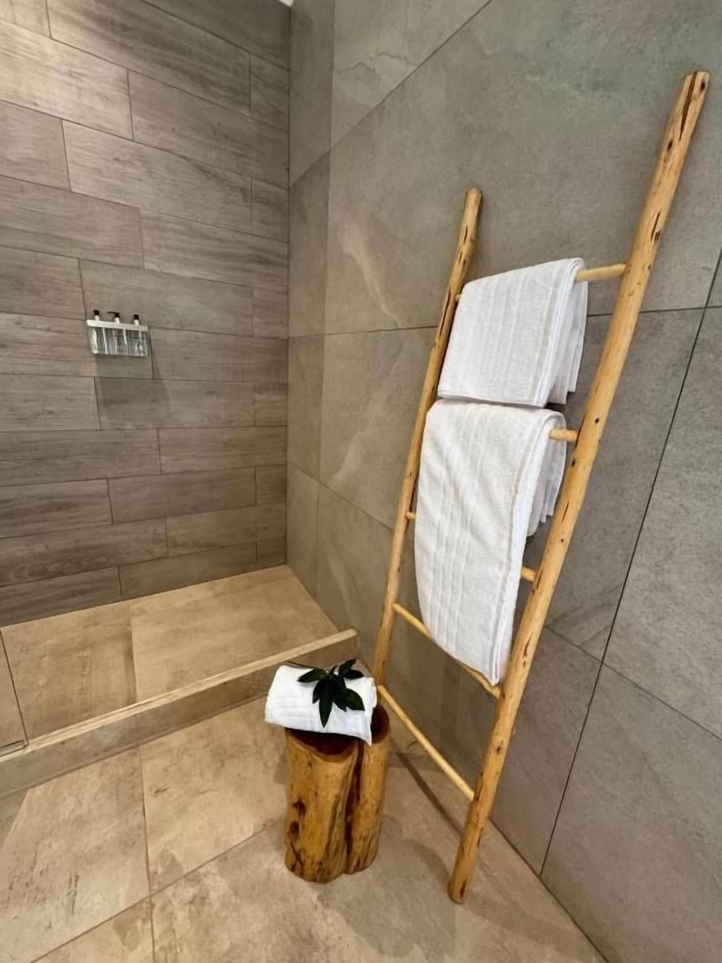 Un espacio de ducha con un toallero de madera que sostiene dos toallas, un taburete pequeño de madera con una toalla y un suelo de baldosas.