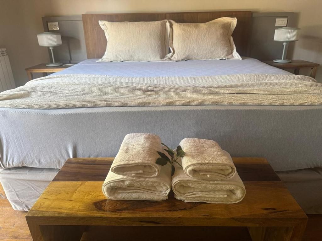 Una cama bien hecha con cubrecama gris y dos almohadas, una mesita de noche con una lámpara, y una pequeña mesa con toallas dobladas al frente.