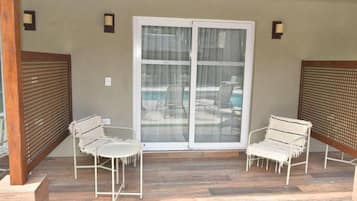 Un patio techado con dos sillas de metal blanco, un vidrio que da a una piscina y una cerca de madera.