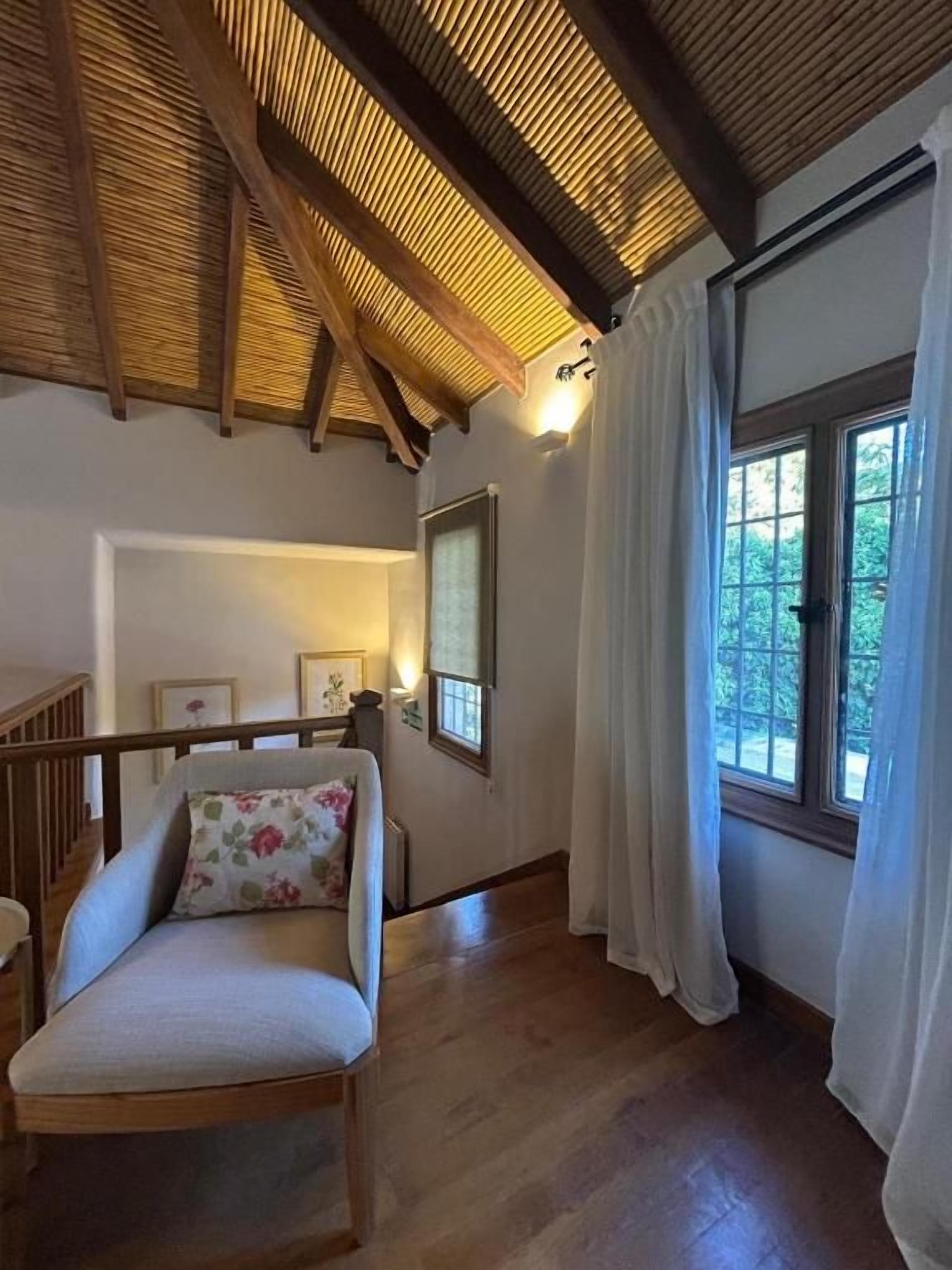 Una habitación con techo de madera, una ventana con cortinas, una silla con cojín y un cuadro en la pared.